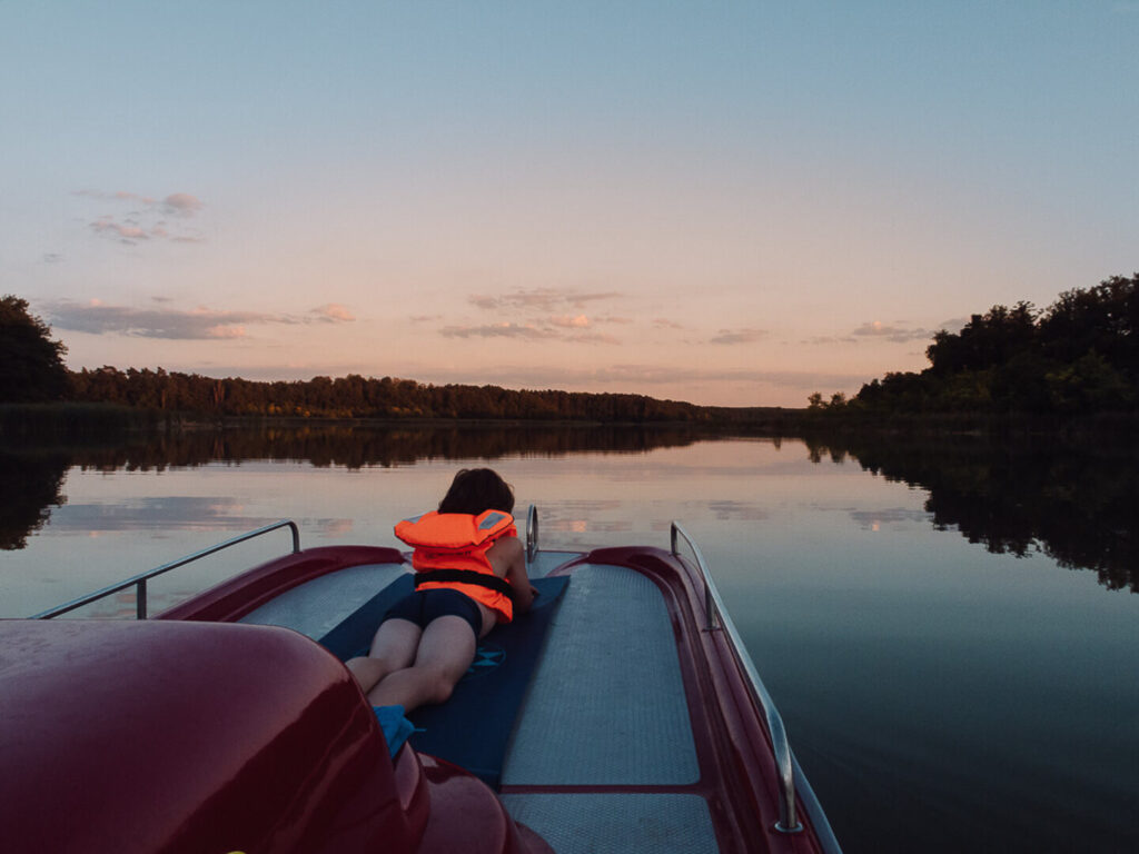 Czerwony rower wodny z platformą na jeziorze, w tle zachód słońca, po lewej i prawej stronie szpalery drzew